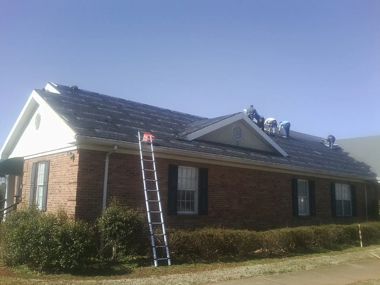 Crew tearing shingles off a brick ranch roof with a ladder set — Metal Roof Repair work by our crew in Evans